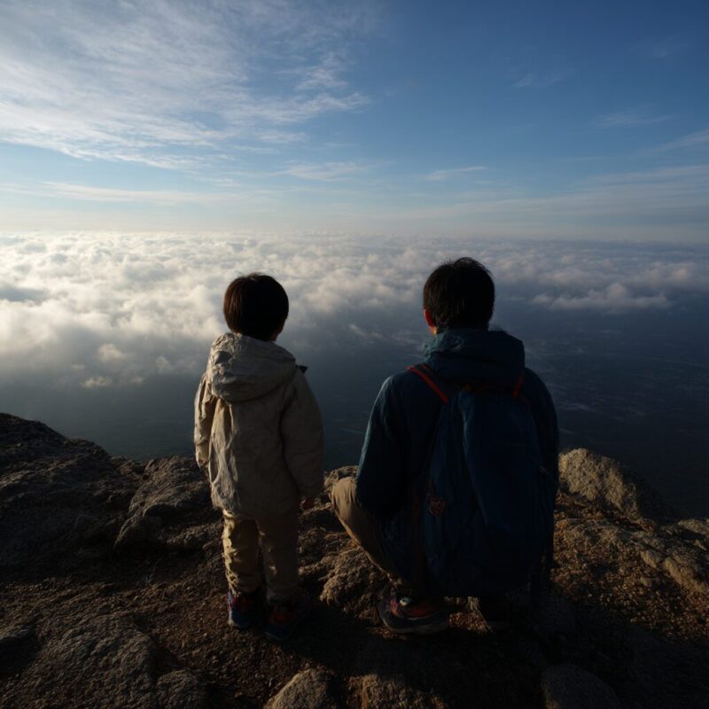 小学2年生の男の子と父親が山頂に立ち、眼下に広がる雲海を見つめている。逆光の中、現代的な登山ファッションで並ぶ後ろ姿が静かに浮かび上がる