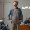 服が山積みになった部屋で、何を着るか悩んでいる40代の男性。シャツの上にTシャツを重ねて鏡の前で考え込んでいる様子。