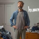 服が山積みになった部屋で、何を着るか悩んでいる40代の男性。シャツの上にTシャツを重ねて鏡の前で考え込んでいる様子。