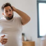 鏡の前でTシャツ姿のお腹を気にしている中年男性。困った表情で頭をかきながら、自分の体型を見つめている。